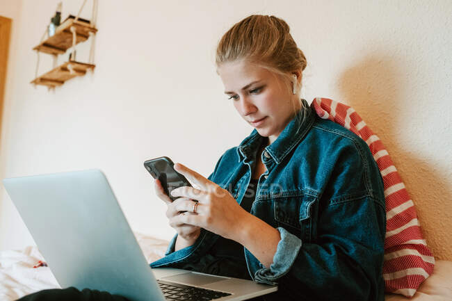 Молода жінка в бездротових навушниках і джинсовій куртці використовує смартфон і ноутбук, розслабляючись на ліжку в сучасній квартирі — стокове фото