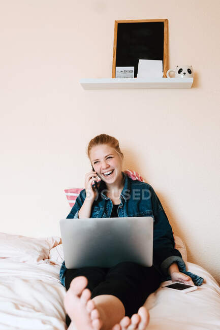 Positivo relaxado jovem fêmea em roupas casuais olhando para longe e rindo enquanto sentado com as pernas cruzadas com laptop em cama macia e fazendo telefonema no quarto acolhedor — Fotografia de Stock