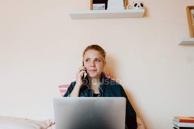 Положительный расслабленной молодой женщины в повседневной одежде глядя в сторону, сидя с ноутбуком на мягкой кровати и сделать телефонный звонок в уютной спальне — стоковое фото