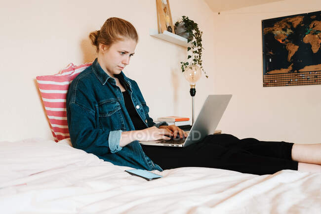 Орієнтована розслаблена молода жінка в повсякденному одязі сидить на м'якому ліжку, використовуючи ноутбук проти світло-бежевої стіни в затишній сучасній квартирі — стокове фото