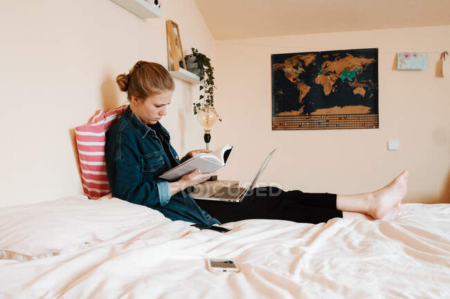 Серьезный задумчивый студентка в повседневной одежде чтение учебника с интересом, сидя со скрещенными ногами на кровати и с помощью ноутбука против современного интерьера светлой уютной квартиры — стоковое фото