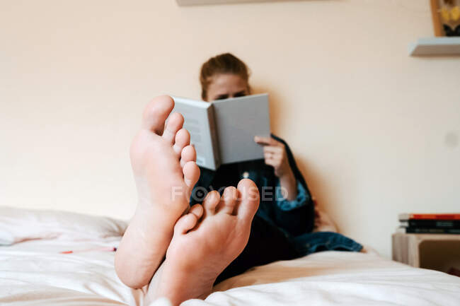 Неузнаваемая босиком женщина в повседневной одежде читает книгу с интересом, расслабляясь в одиночестве на мягкой кровати оставаться в безопасности дома — стоковое фото