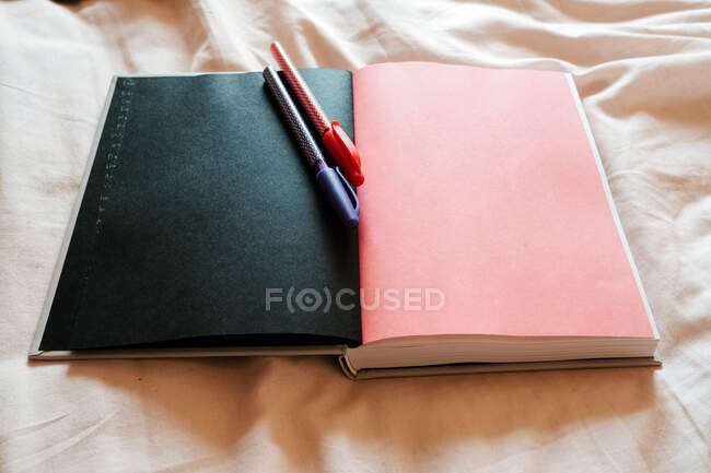 Do caderno acima aberto com páginas pretas e rosa em branco na composição com canetas vermelhas e azuis no edredão branco na cama no quarto leve do apartamento moderno — Fotografia de Stock
