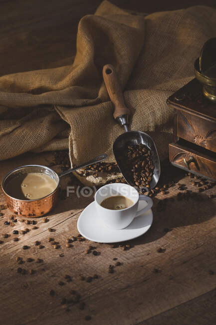 Frischer schwarzer Kaffee in weißer Keramiktasse auf Untertasse in der Nähe von Kaffeemühle und Kaffeebohnen auf Holztisch — Stockfoto
