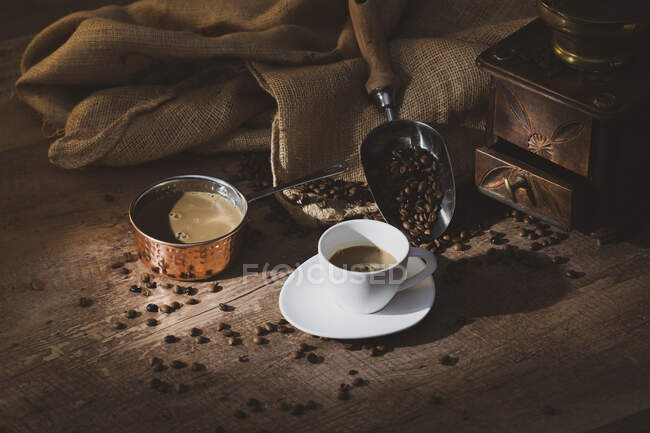 Frischer schwarzer Kaffee in weißer Keramiktasse auf Untertasse in der Nähe von Kaffeemühle und Kaffeebohnen auf Holztisch — Stockfoto