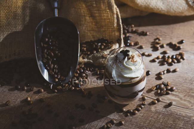 Copa de postre dulce con chocolate y café adornado con crema colocada en mesa de madera cerca de cucharada de metal con granos de café - foto de stock