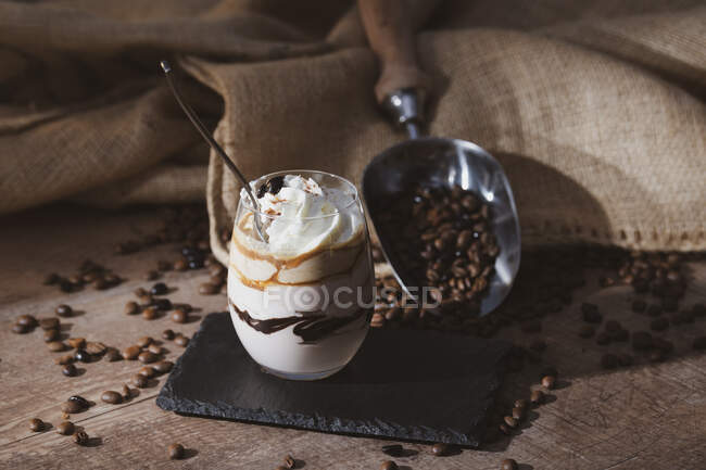 Glass of sweet dessert with chocolate and coffee garnished with cream placed on wooden table near metal scoop with coffee beans — Stock Photo