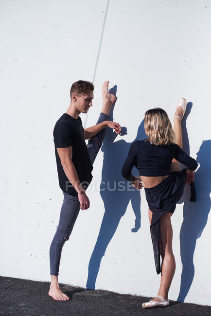 Volle Länge des flexiblen Paares, das einander ansieht und mit erhobenem Bein auf weißer Wand mit fallendem Schatten an sonnigen Tagen teilt — Stockfoto