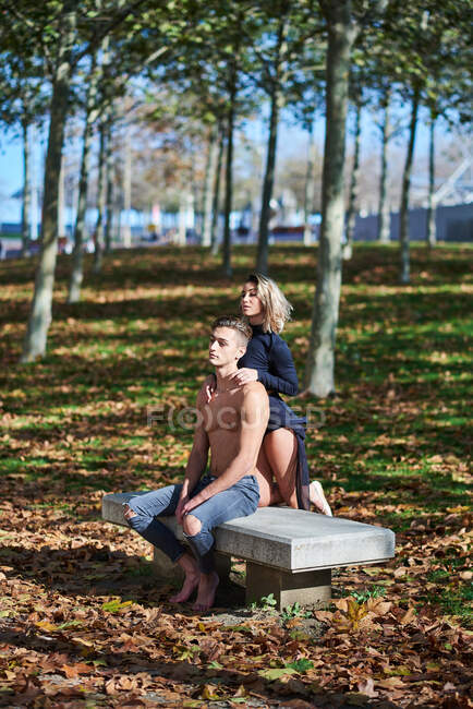 Jeune homme en forme torse nu assis sur le banc dans le parc tandis que la grâce attrayante dame debout derrière et mettre la main sur les épaules des gars dans le parc d'automne — Photo de stock