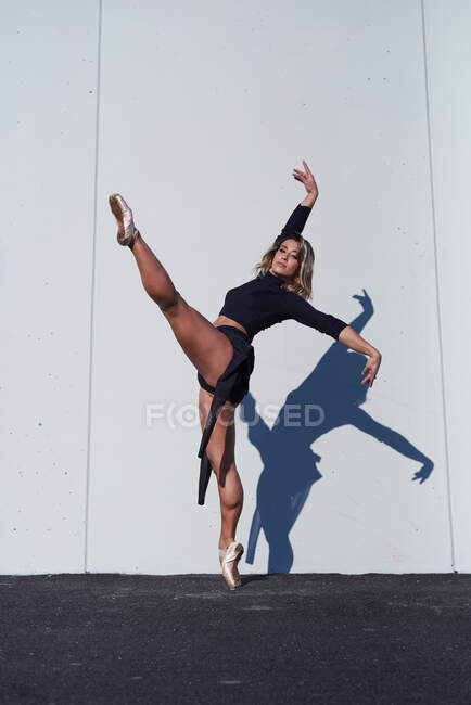 Full body of feminine dancer in black costume and pointe shoes performing posture looking at camera while standing on tiptoe on one leg against white wall with falling shadow — Stock Photo