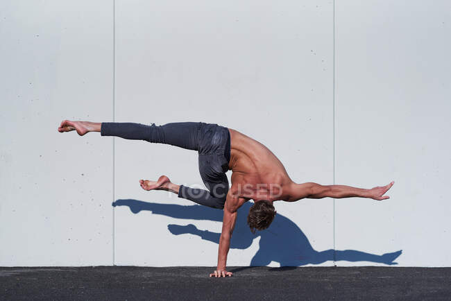Back view of faceless muscular shirtless acrobat standing on one hand while stretching one leg perpendicular to floor and other bending at knee during training against white wall — Stock Photo