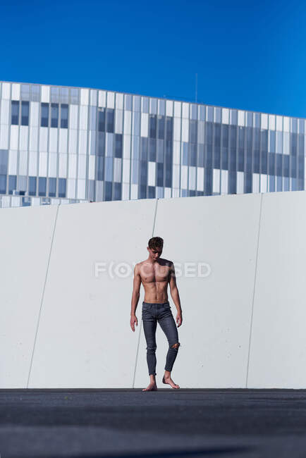 Sportif sans chemise se reposant après la performance debout contre un mur avec un ciel bleu clair sur le toit du bâtiment moderne — Photo de stock