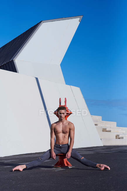 Handsome athlete with naked torso sitting on iron structure on roof of building with legs spread wide and outstretched toes and looking at camera — Stock Photo