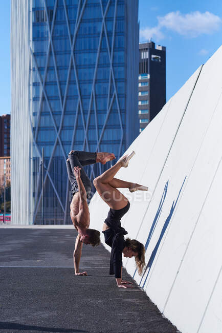 Vista lateral del atleta y la mujer del hombre sin camisa del ajuste que realizan el handstand contra la pared en la azotea contra el rascacielos moderno en día soleado - foto de stock