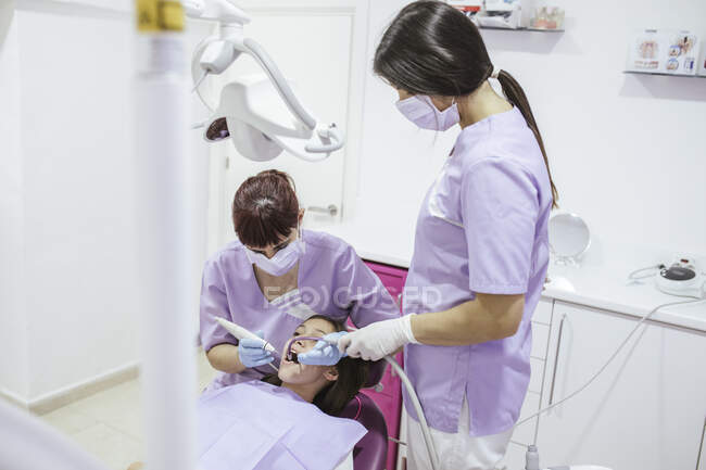 Женщина-стоматолог в форме и маске вылечивает зубы пациентки с ассистенткой в современной стоматологической клинике — стоковое фото