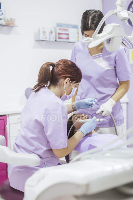 Жінка-стоматолог в уніформі та масці для лікування зубів пацієнта з помічницею жінки в сучасній стоматологічній клініці — стокове фото