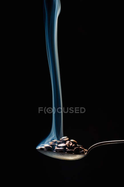 Closeup metal spoon with fresh coffee grains and white steam flowing up in dark room on black background — Stock Photo