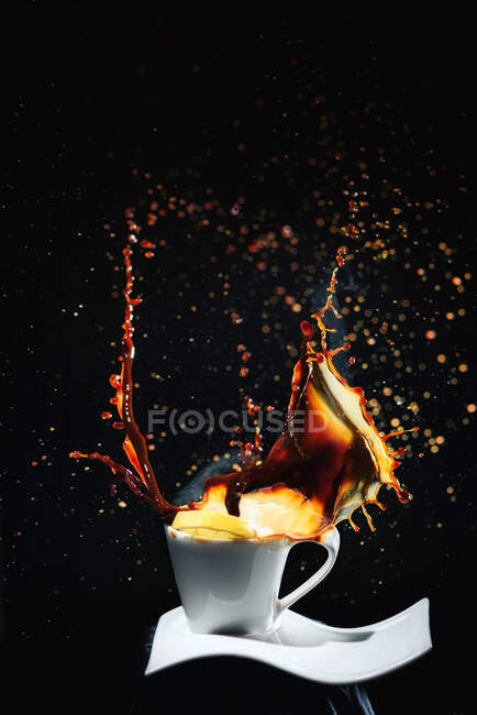 Fallende Scheibe frischer Zitrone in spritzenden heißen frischen Kaffee in weißer Tasse auf weißem Teller in dunklen Raum auf schwarzem Hintergrund — Stockfoto