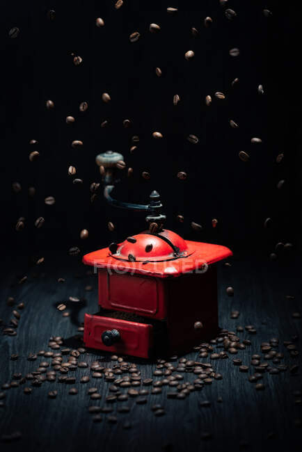 Vintage metal red coffee grinder placed on black wooden surface and falling coffee grains in dark room on black background — Stock Photo