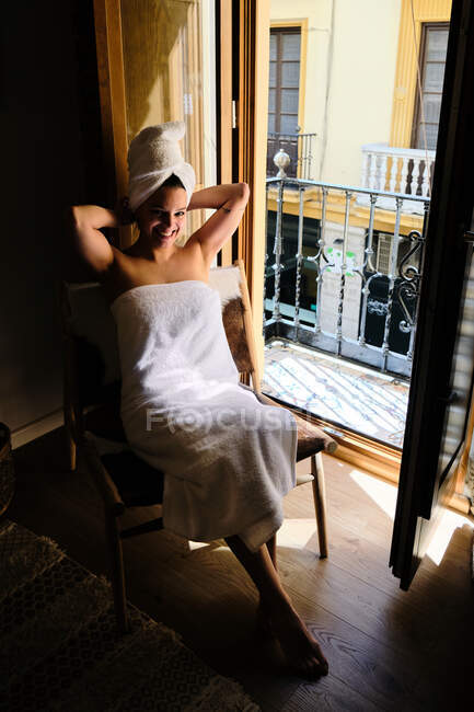 Donna felice in asciugamano bianco sul corpo e la testa seduta sulla sedia dalla porta del balcone appoggiato sulle mani in appartamento con pavimento in legno nella giornata di sole e guardando la fotocamera — Foto stock