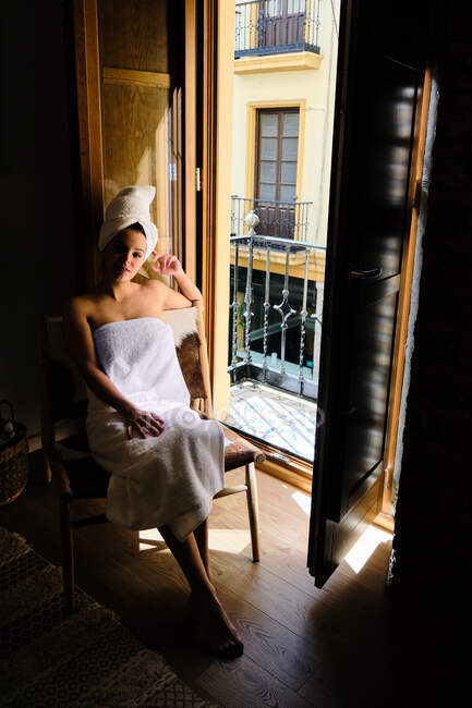 Серен расслабленная женщина в белом плаще на теле и голове, сидящая на стуле у балконной двери в квартире с деревянным полом в солнечный день и смотрящая в камеру — стоковое фото
