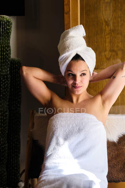 Mulher feliz em toalha branca no corpo e cabeça sentada na cadeira pela porta apoiada nas mãos em apartamento no dia ensolarado e olhando para longe — Fotografia de Stock
