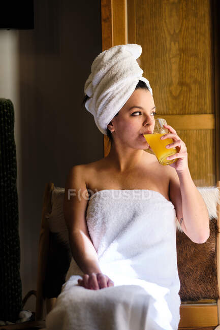 Nachdenkliche ruhige junge Frau nach der Dusche in Handtücher gehüllt, genießt frischen Saft, während sie neben der Holztür sitzt und an sonnigen Tagen wegschaut — Stockfoto