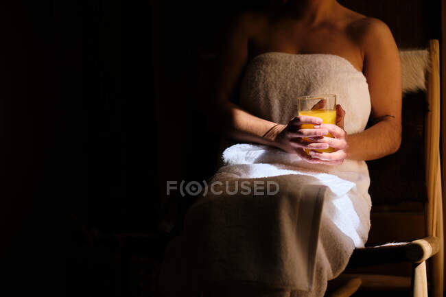 Ritagliato femmina calma irriconoscibile avvolto in asciugamani dopo la doccia godendo di succo fresco mentre seduto vicino alla porta di legno e guardando lontano nella giornata di sole — Foto stock