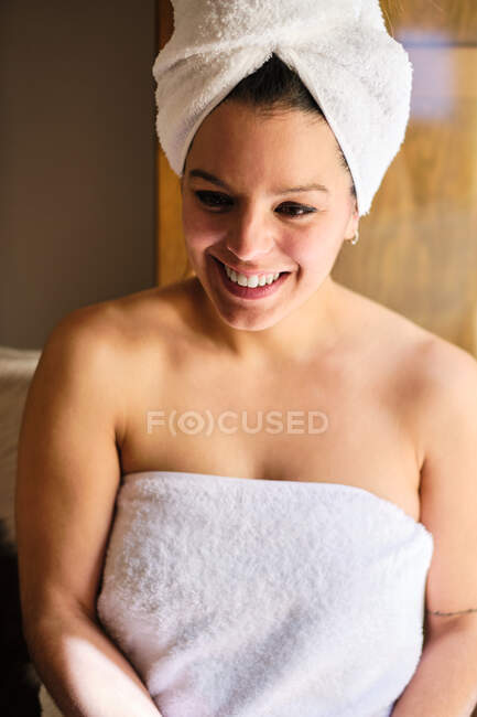 Happy female in white towel on body and head sitting on chair at home on sunny day and looking away — Stock Photo