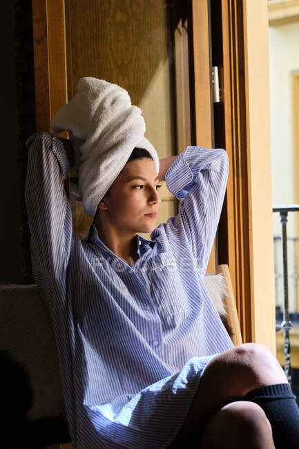 Pensive Frau in lässigem Outfit mit Handtuch auf nassem Haar sitzt mit überkreuzten Beinen und Händen hinter dem Kopf in der Nähe einer Holztür im Sonnenlicht und schaut weg — Stockfoto