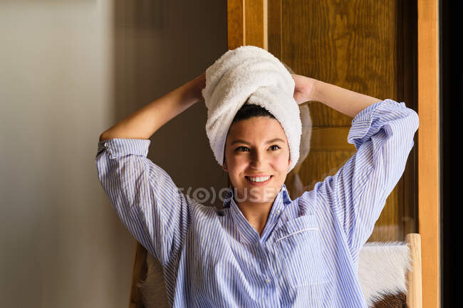 Усміхнена жінка з білим махровим рушником на голові стоїть нахилившись на руку і дивлячись на камеру в квартирі в сонячний день — стокове фото