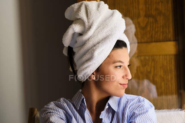 Улыбающаяся женщина с белым махровым полотенцем на голове сидит на стуле и смотрит на квартиру в солнечный день — стоковое фото