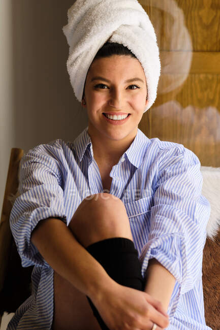 Mulher sorridente com toalha branca na cabeça sentada em uma cadeira olhando para a câmera no apartamento no dia ensolarado — Fotografia de Stock