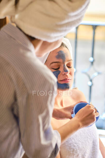 Anonyme weibliche Anwendung blauer Tonmaske auf das Gesicht einer gelassenen Frau mit geschlossenen Augen in weißem Handtuch während des Eingriffs zu Hause — Stockfoto