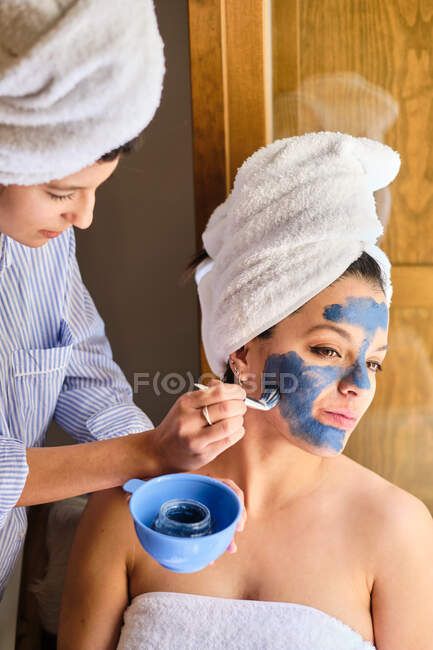 Женщина надевает синюю глиняную маску на лицо безмятежной подруги, отводя взгляд с полотенцем, завернутым в голову после ванны, сидя рядом с балконом под солнечным светом — стоковое фото