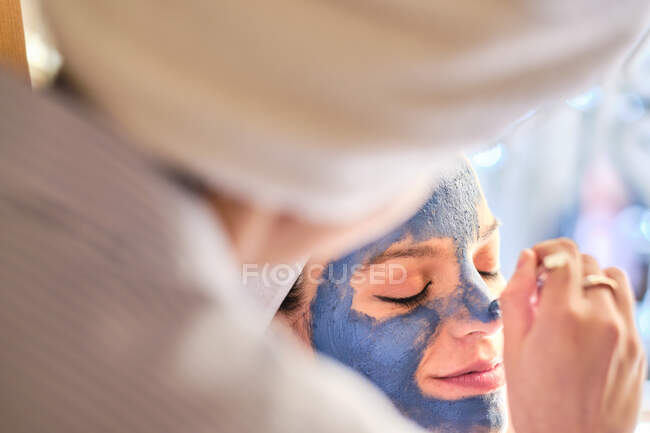 Visão traseira da fêmea anônima aplicando máscara de barro azul no rosto de mulher serena com olhos fechados em toalha branca durante o procedimento em casa — Fotografia de Stock