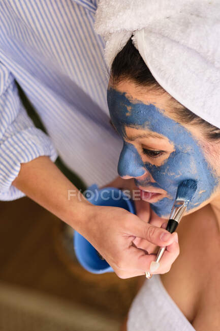 Анонимная женщина надевает синюю глиняную маску на лицо безмятежной женщины, смотрящей в белое полотенце во время процедуры дома — стоковое фото
