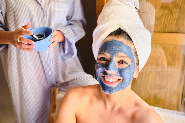 Anonyme Frau trägt blaue Lehmmaske auf Gesicht einer fröhlichen Frau auf, die in weißes Handtuch während der Behandlung zu Hause in die Kamera schaut — Stockfoto