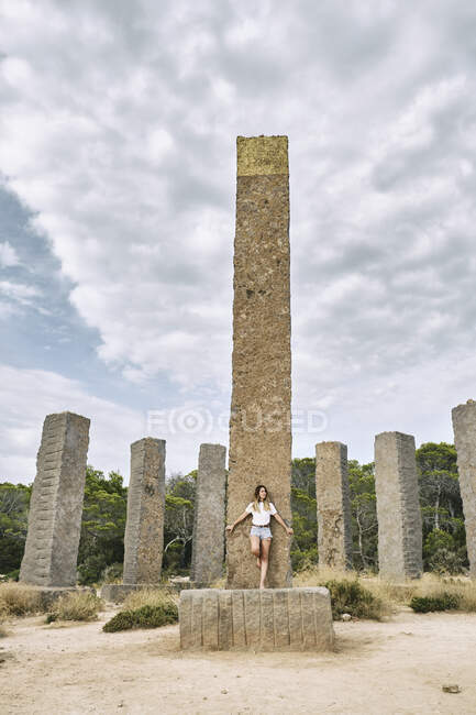 Viajante feminino de corpo inteiro em roupas casuais de verão em colunas de pedra velhas e olhando para longe durante as férias em Ibiza ensolarado — Fotografia de Stock