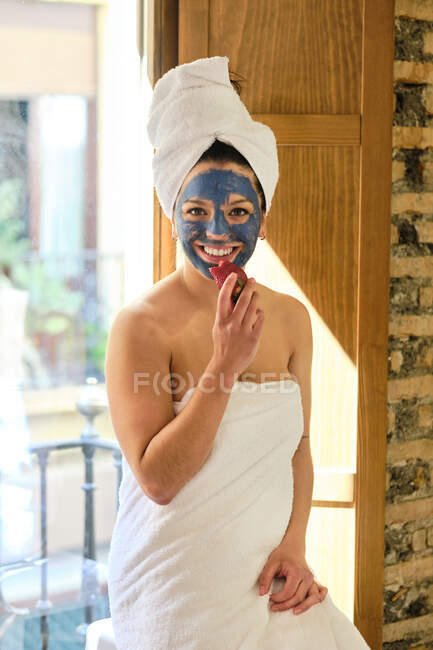 Cheerful female in white soft towel with blue clay mask applied on face standing near door while eating strawberry and looking at camera in sunlight — Stock Photo