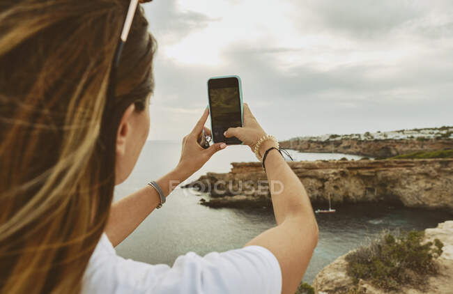 Back view of anonymous female tourist in casual clothes and sunglasses taking pictures on smartphone while standing on rocky cliff on seashore on cloudy day in Ibiza — Stock Photo