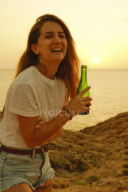 Lächelnde junge Frau mit einer Flasche Bier während des Sonnenuntergangs am Strand — Stockfoto