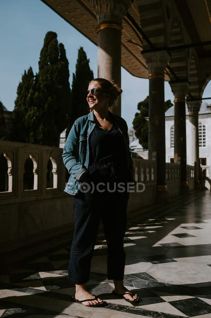 Full body of cheerful female tourist in casual clothes and sunglasses standing on terrace of old stone oriental building holding hands in pockets and looking away while enjoying warm summer evening — Stock Photo