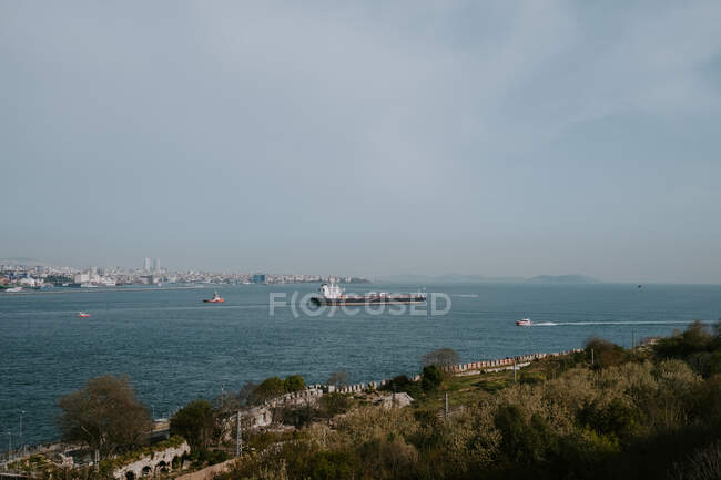 Majestic panoramic scenery of shore with trees against calm sea water with distant sailing boats coastal city and blue sky on summer day — Stock Photo