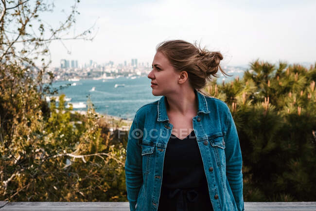 Pensive young female tourist in casual jeans jacket and black dress standing near green trees against amazing view of blue sea and cloudy sky in Istanbul and looking away — Stock Photo