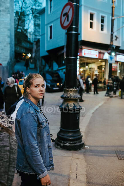Vista lateral da jovem senhora loira em roupa casual em pé na rua e olhando para a câmera enquanto se prepara cruzando rua — Fotografia de Stock