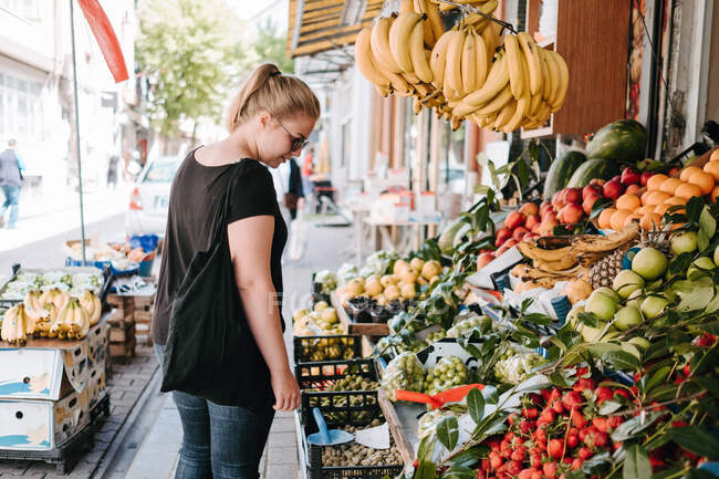 Вид позитивной женщины в повседневной одежде и солнцезащитных очках, стоящей возле фруктового прилавка на турецком рынке и торгующей товарами во время прогулки по улицам города — стоковое фото