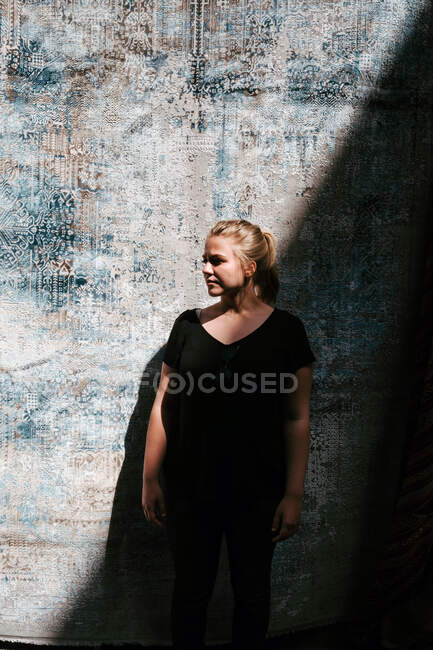 Jovem mulher positiva com cabelo loiro em roupa preta casual em pé contra tapete turco tecido e olhando para longe no dia ensolarado — Fotografia de Stock