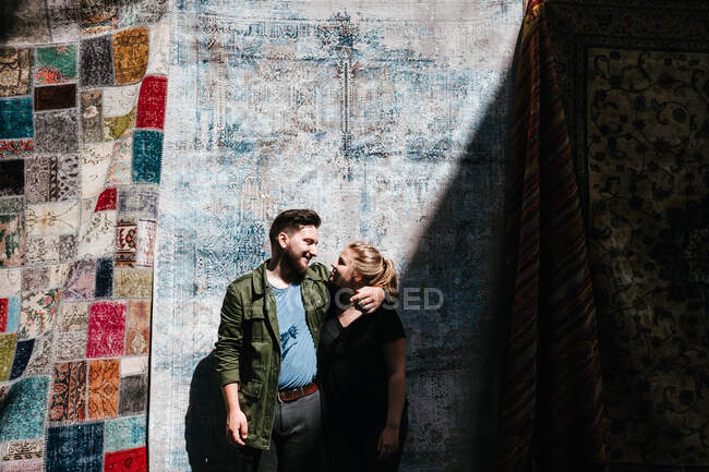 Alegre pareja sonriente abrazándose mirando el uno al otro y de pie sobre el fondo de alfombras turcas ornamentales en el mercado en Estambul durante el día - foto de stock
