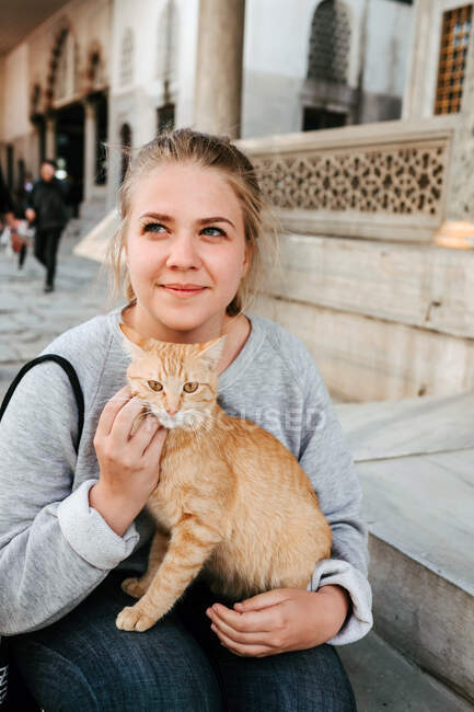 Fröhliche junge Touristin in lässiger Kleidung mit roter Katze in der Hand und Streichelkatze im Gesicht, während sie in Istanbul auf der Straße sitzt und wegschaut — Stockfoto